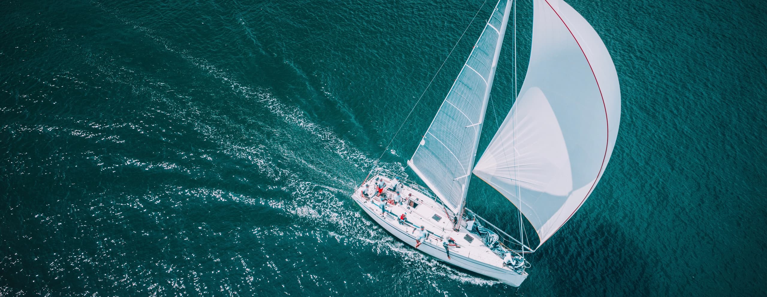 An overhead birds-eye view of a white sailboat sailing through deep blue water, leaving a white wake behind it.