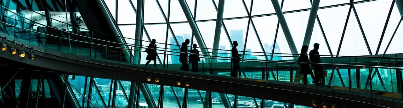 Silhouettes of business professionals walking along a modern glass-enclosed indoor bridge in an office building.