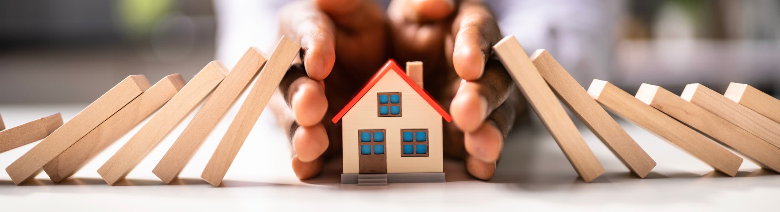 Hands cupping and protecting a small model house from a falling line of wooden dominoes, representing real estate protection and insurance.