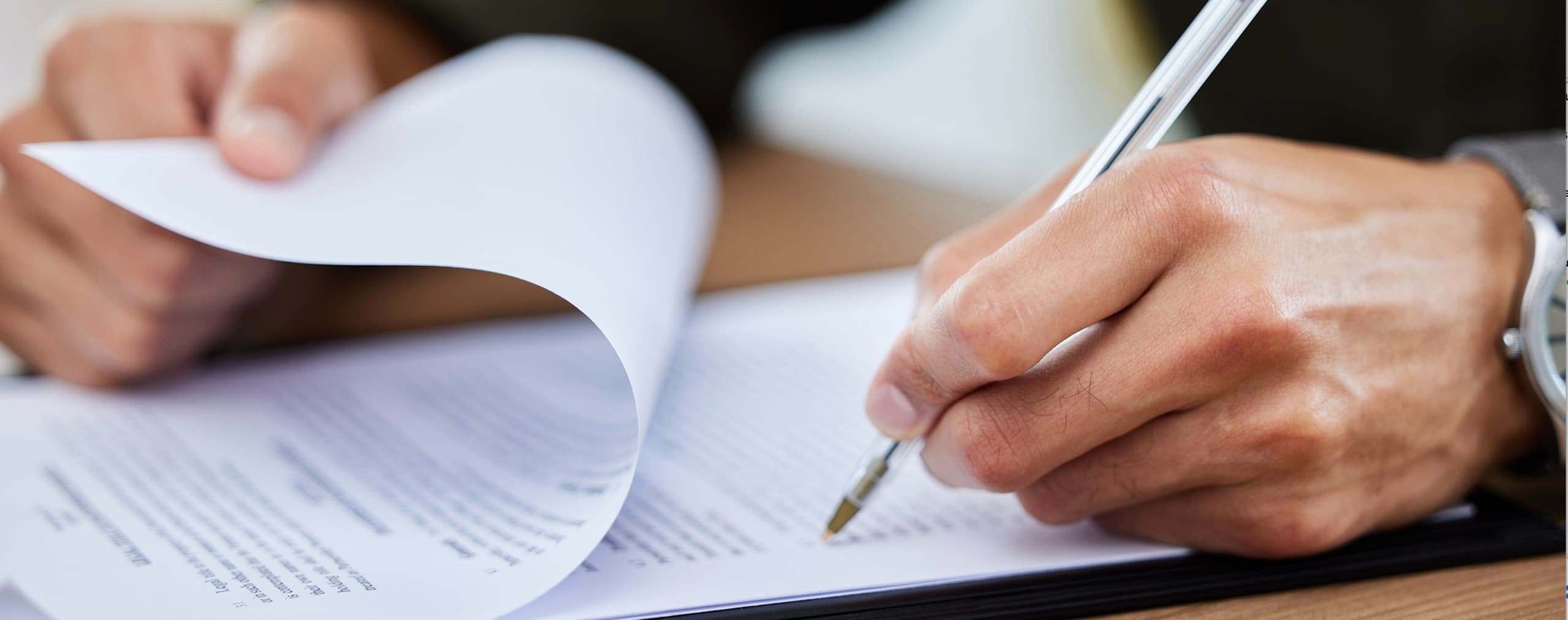 Close-up of a person's hands reviewing and signing a paper document with a pen on a desk.