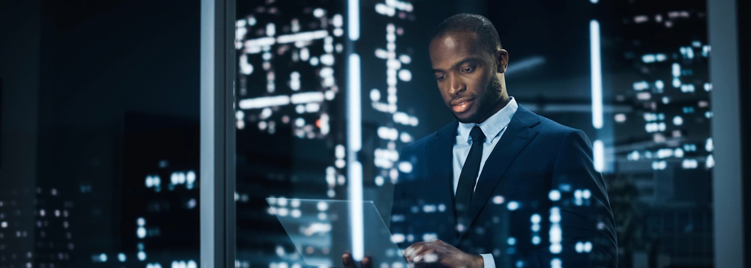Businessman standing with a laptop in his hand, in front of a window with the outside building lights reflecting off of the window.
