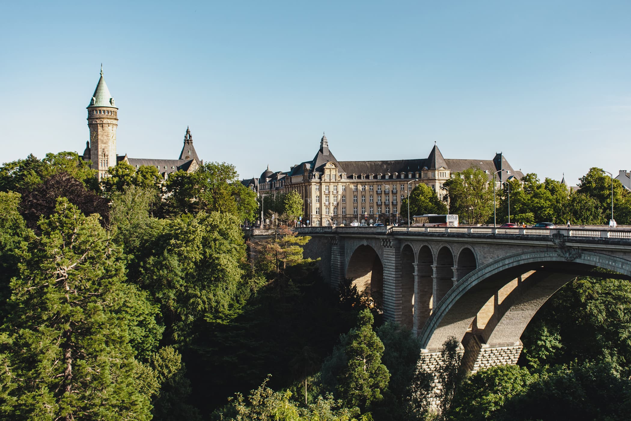 The historic Adolphe Bridge spanning a lush green valley in Luxembourg City, with the State Savings Bank tower in the background.