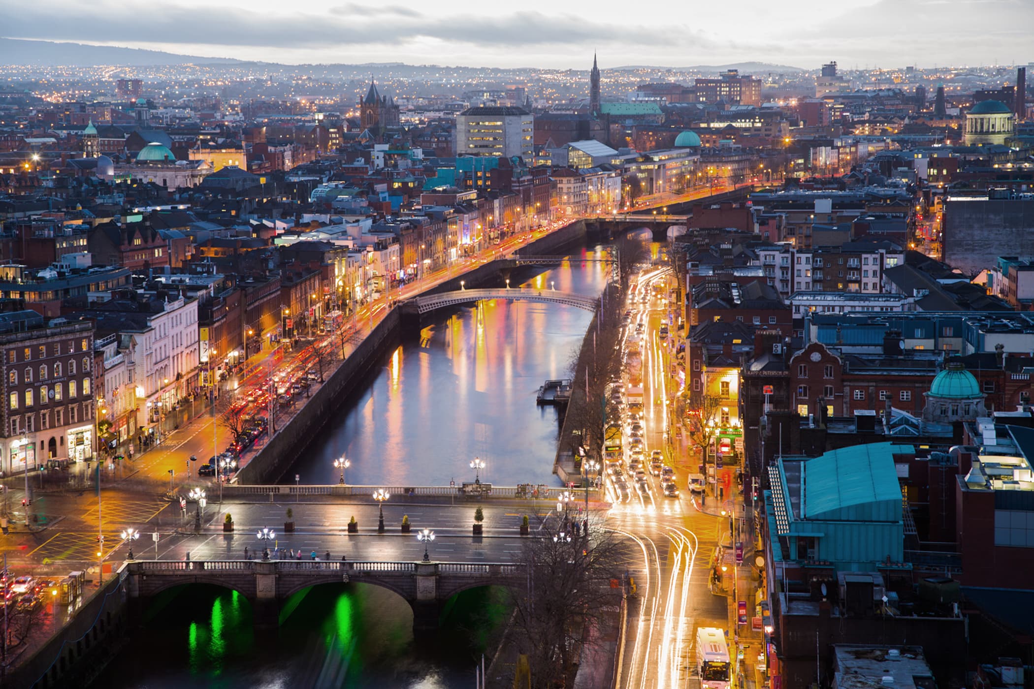 A twilight cityscape of Dublin, Ireland, with street lights and traffic trails reflecting on the River Liffey.