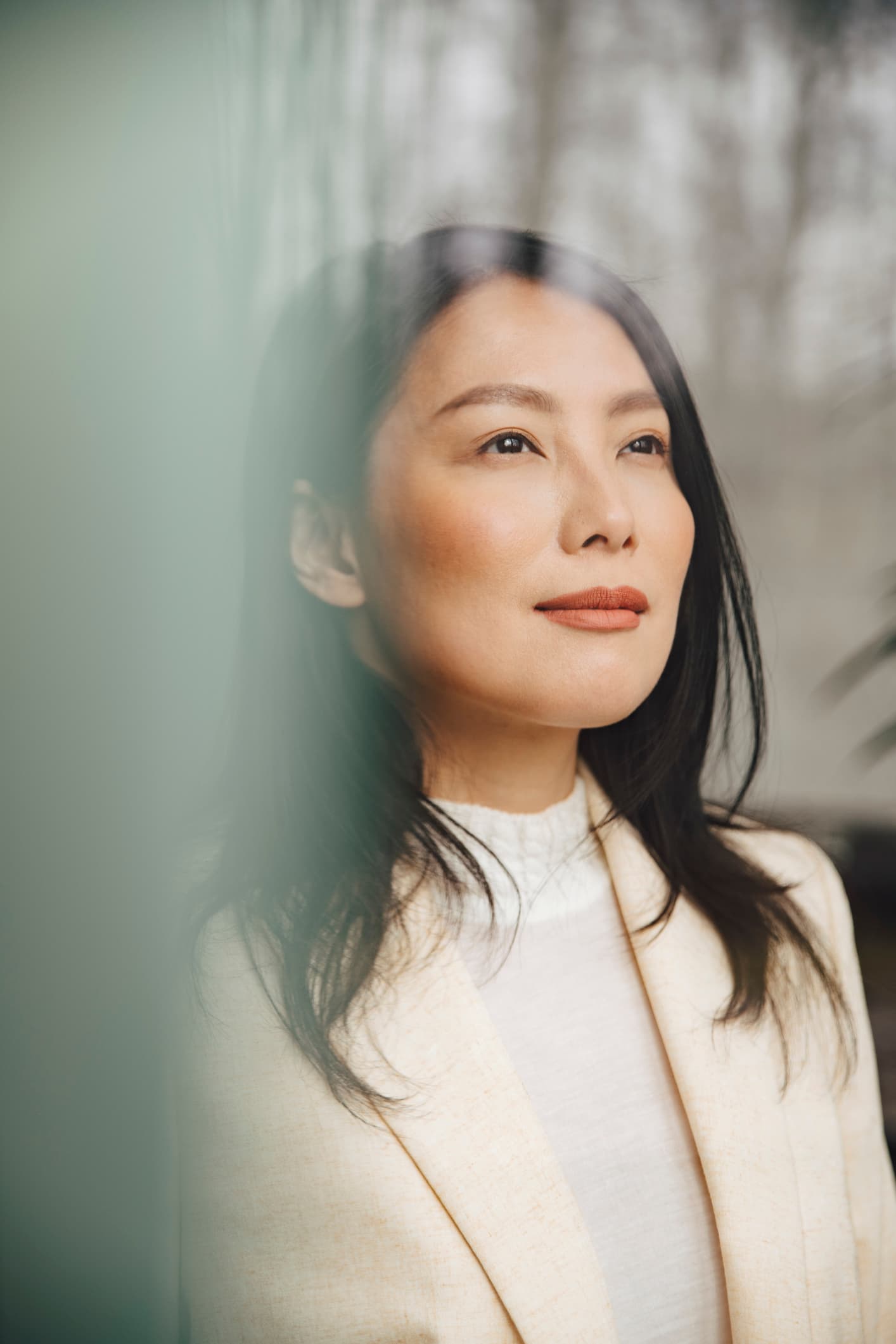 A thoughtful East Asian woman with dark hair, wearing a white blazer, looking off-camera next to a glass window.