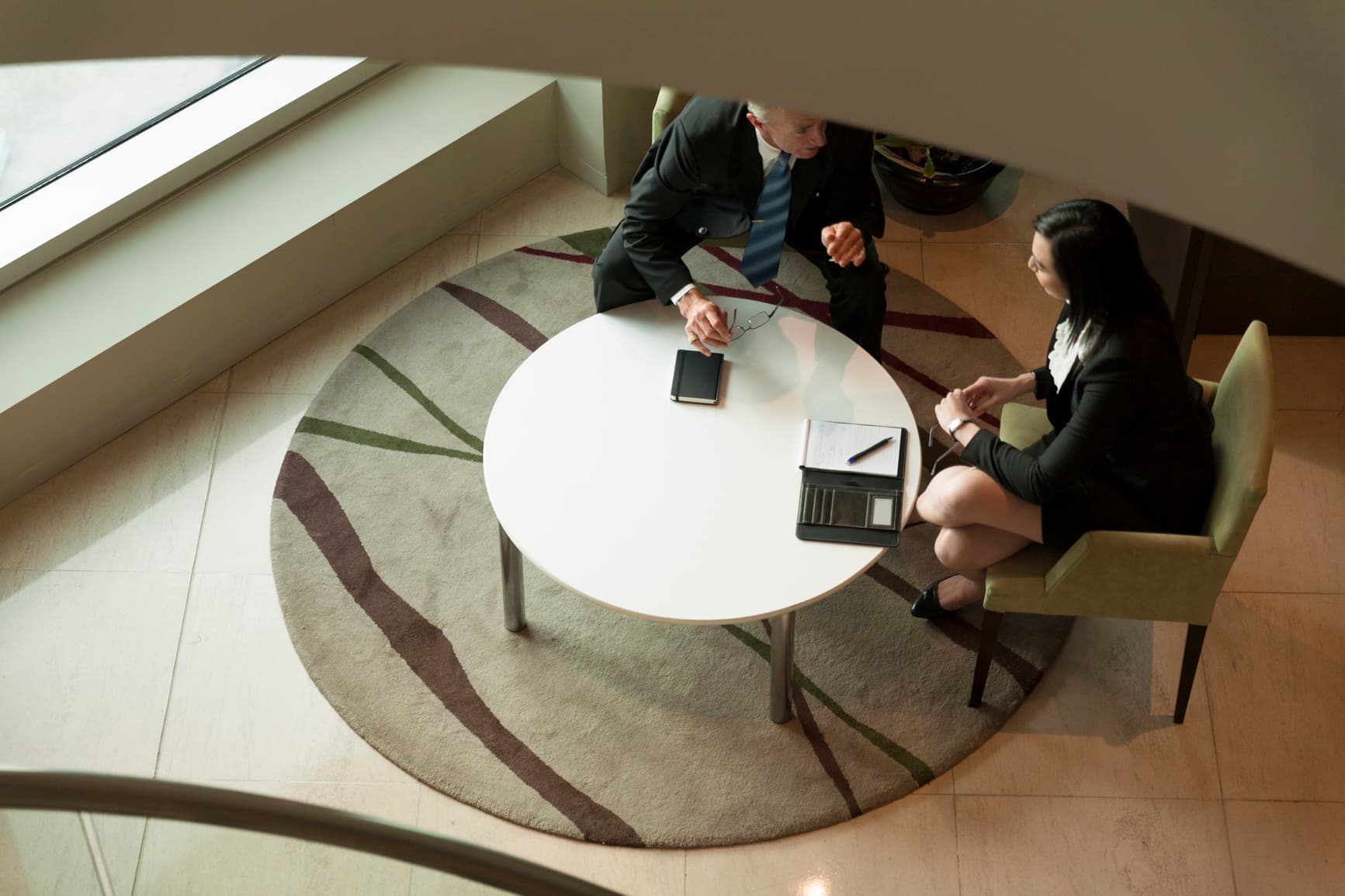 A high-angle, birds-eye view of a businessman and a businesswoman in suits discussing a project around a round white table.