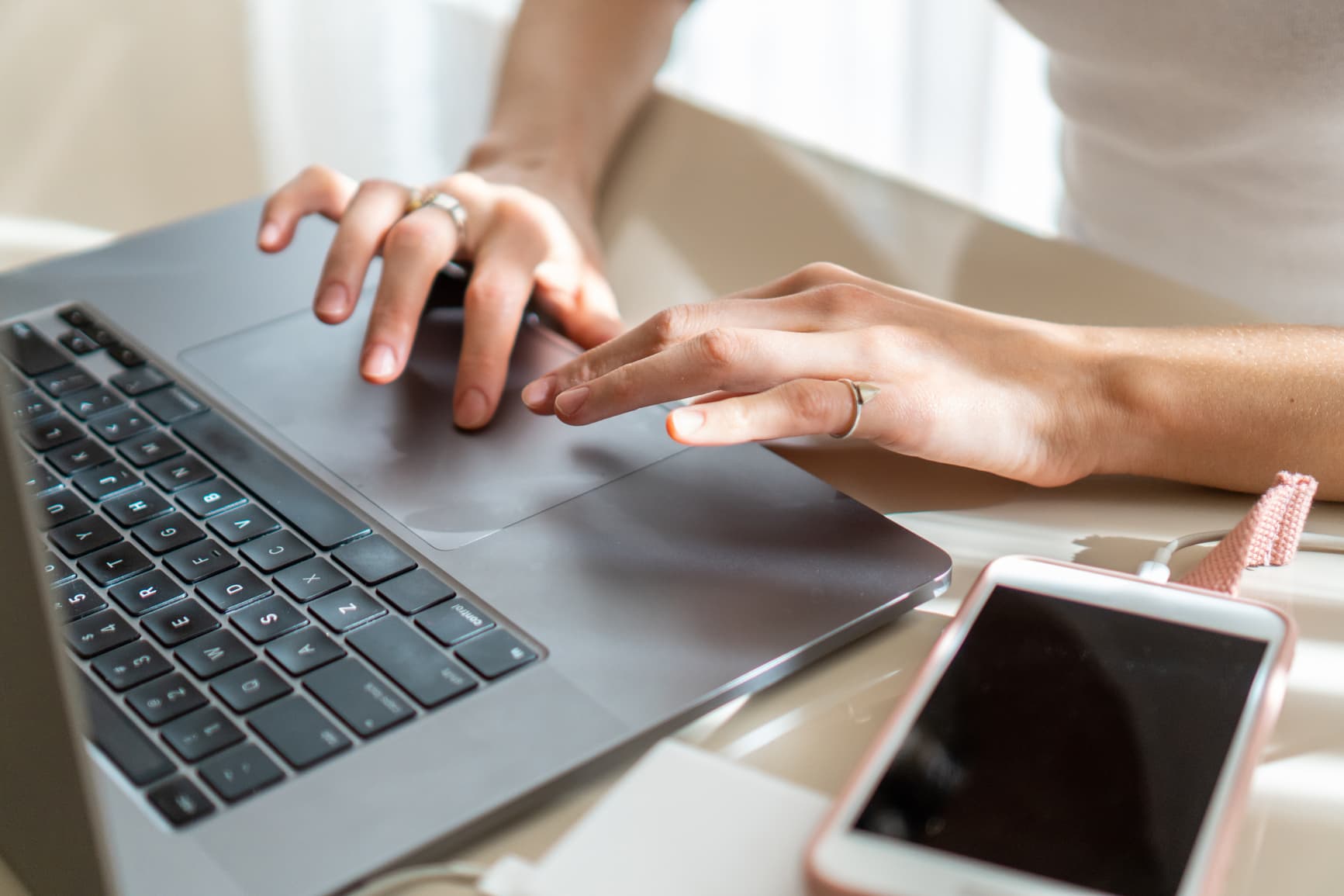 A close-up of a person's hands using a laptop trackpad and keyboard next to a smartphone.