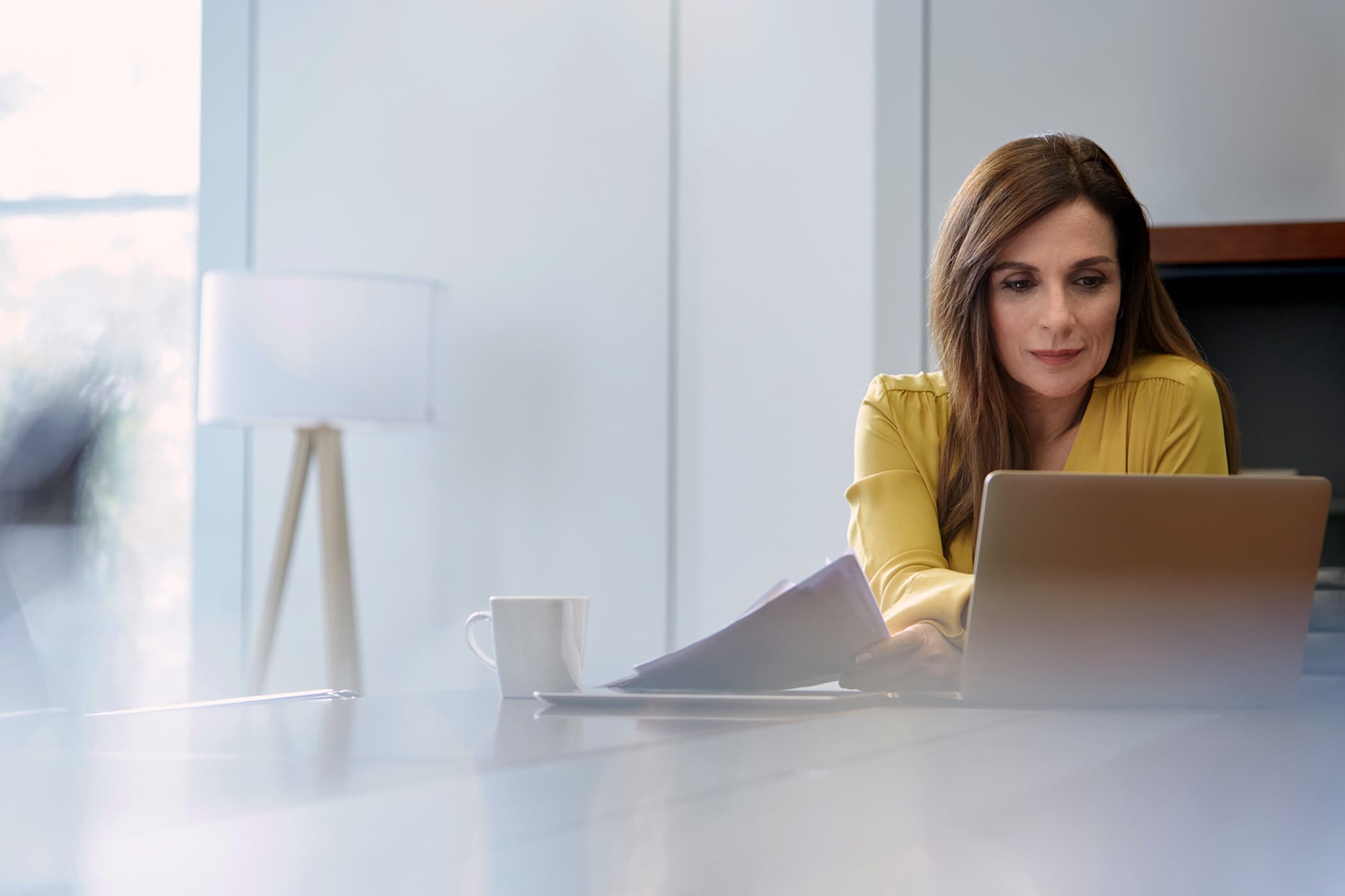 A woman in a yellow blouse sitting at a white desk, holding documents and looking at her laptop screen.