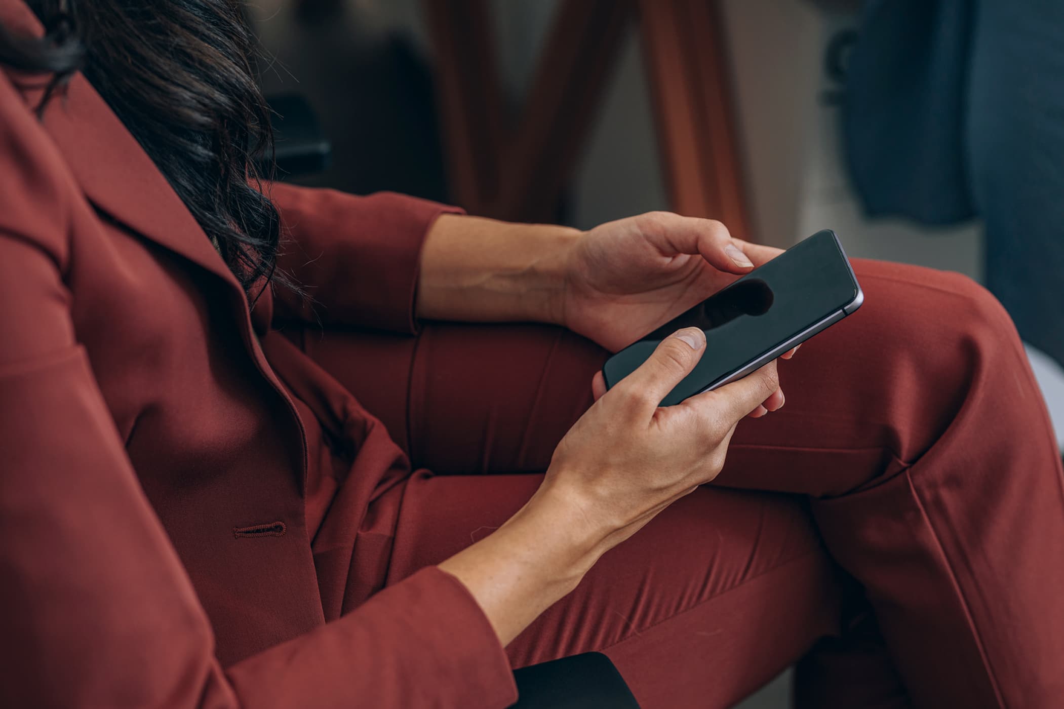 A close-up of a person in a rust-colored suit sitting and holding a smartphone with both hands.
