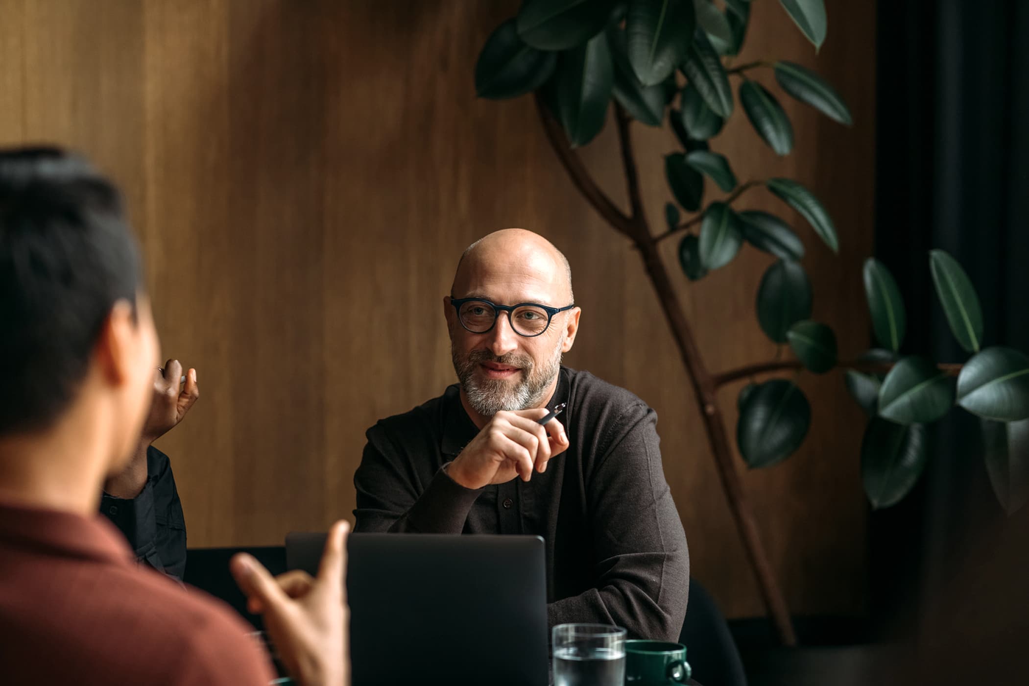 A bald man with glasses and a beard smiling while listening to a colleague speak during a meeting.
