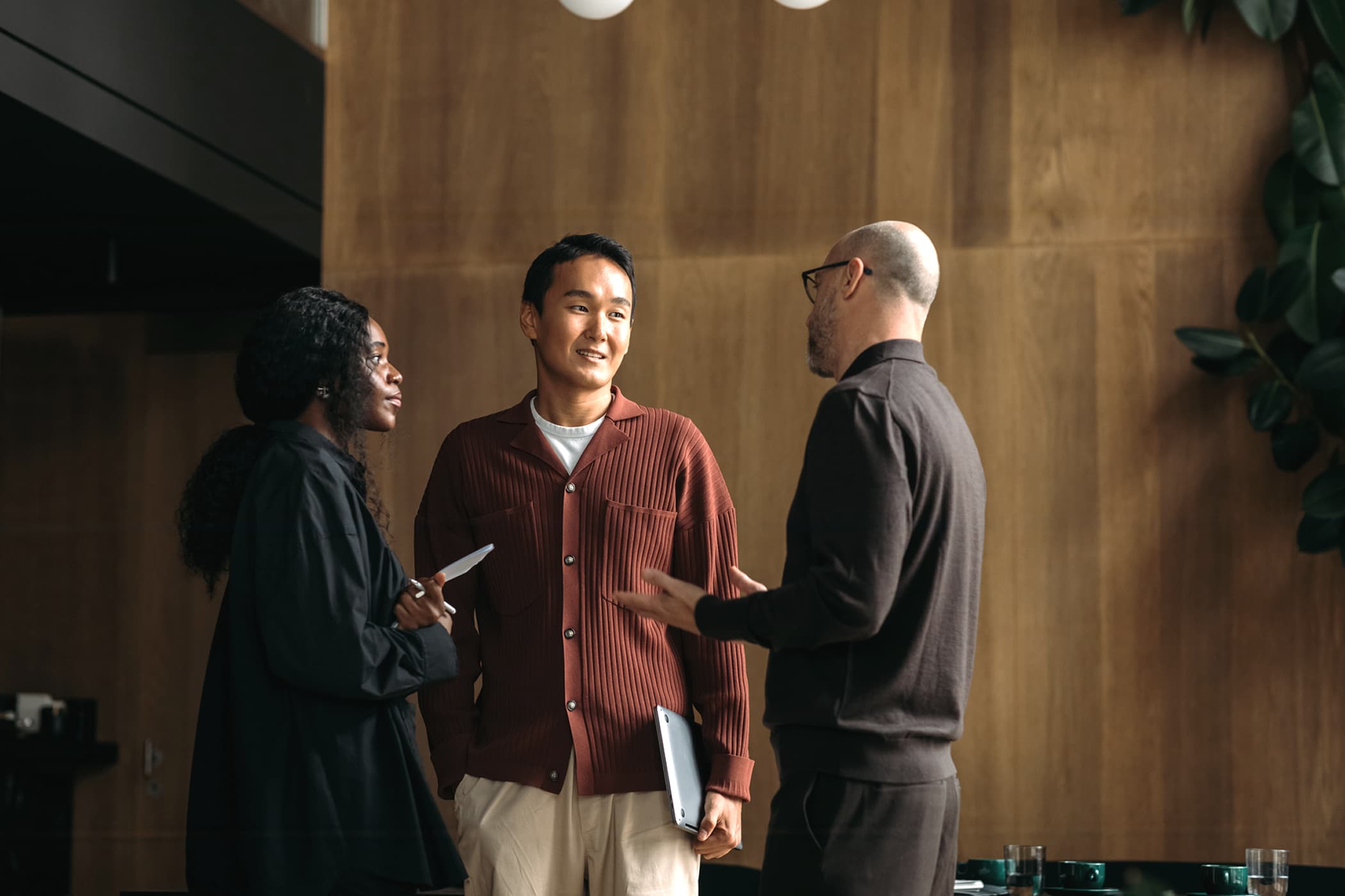 Three diverse colleagues talking in front of a wooden wall in a modern office lobby.