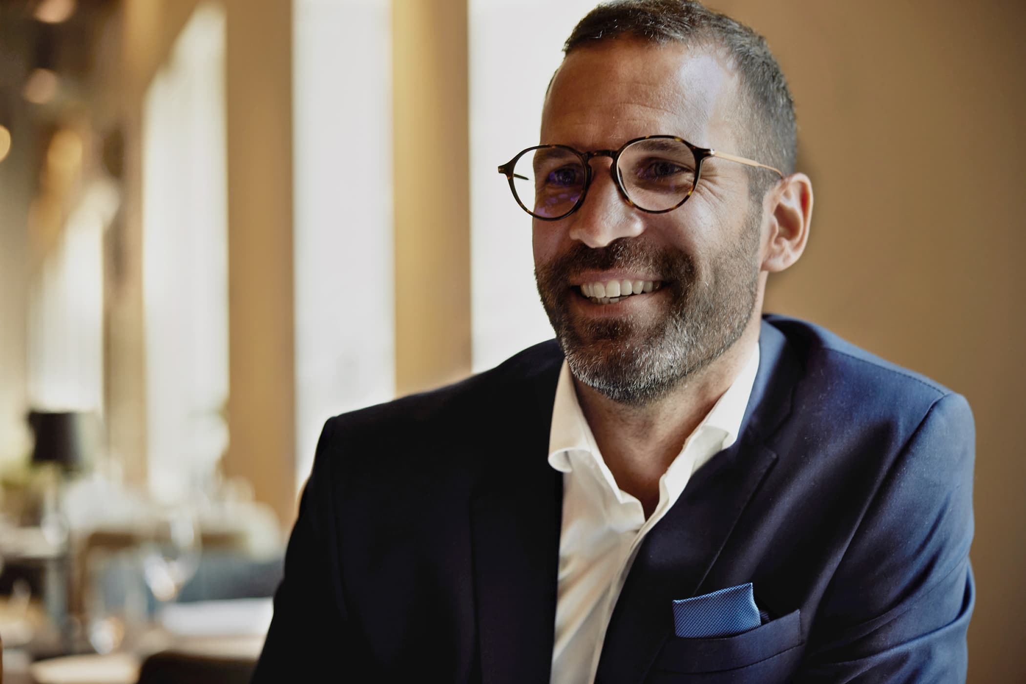 A smiling man with a beard, wearing glasses and a navy blue suit with a white shirt and pocket square.