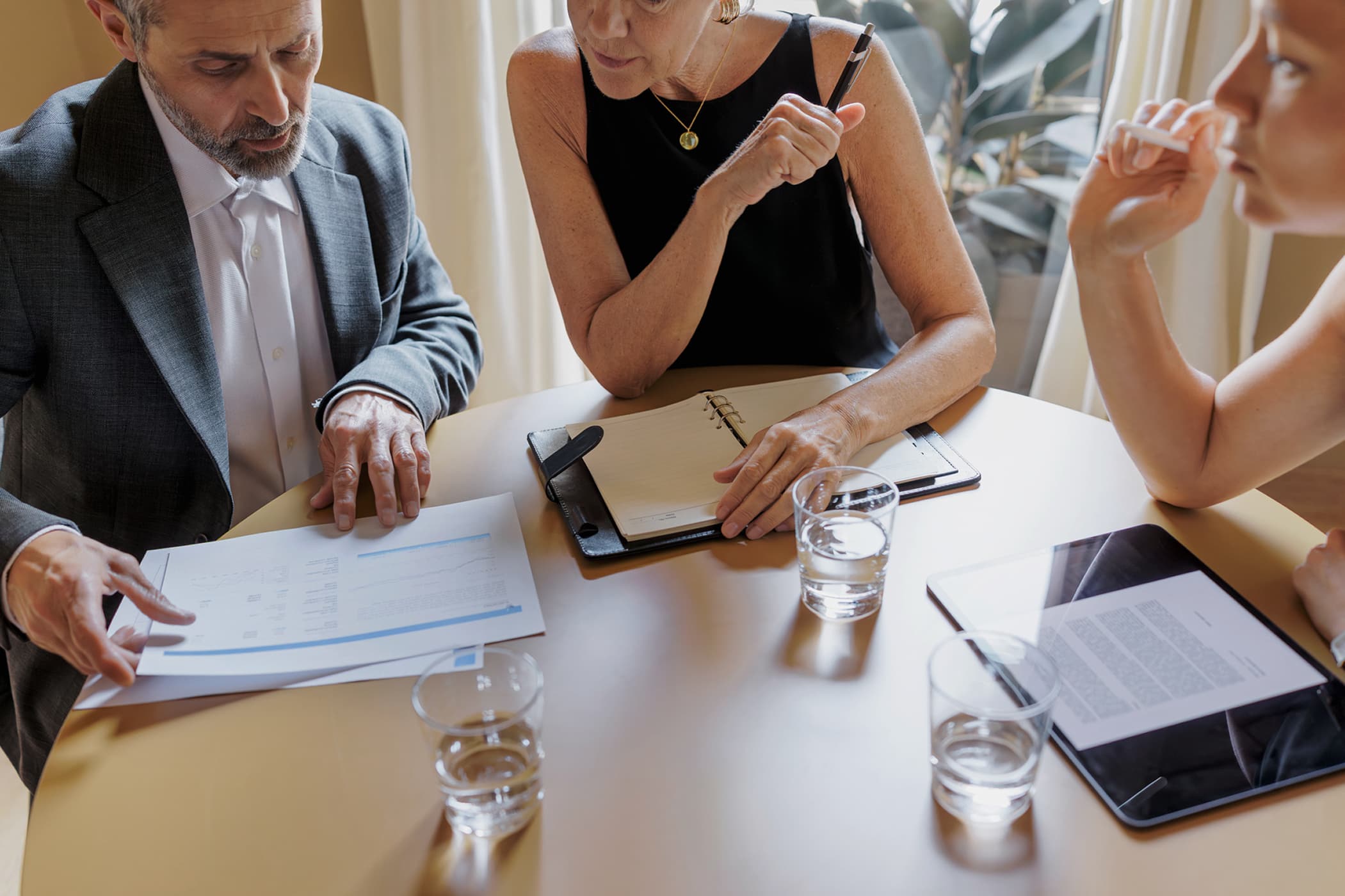 A close-up of three business professionals sitting at a round table, reviewing a printed document and a digital tablet.