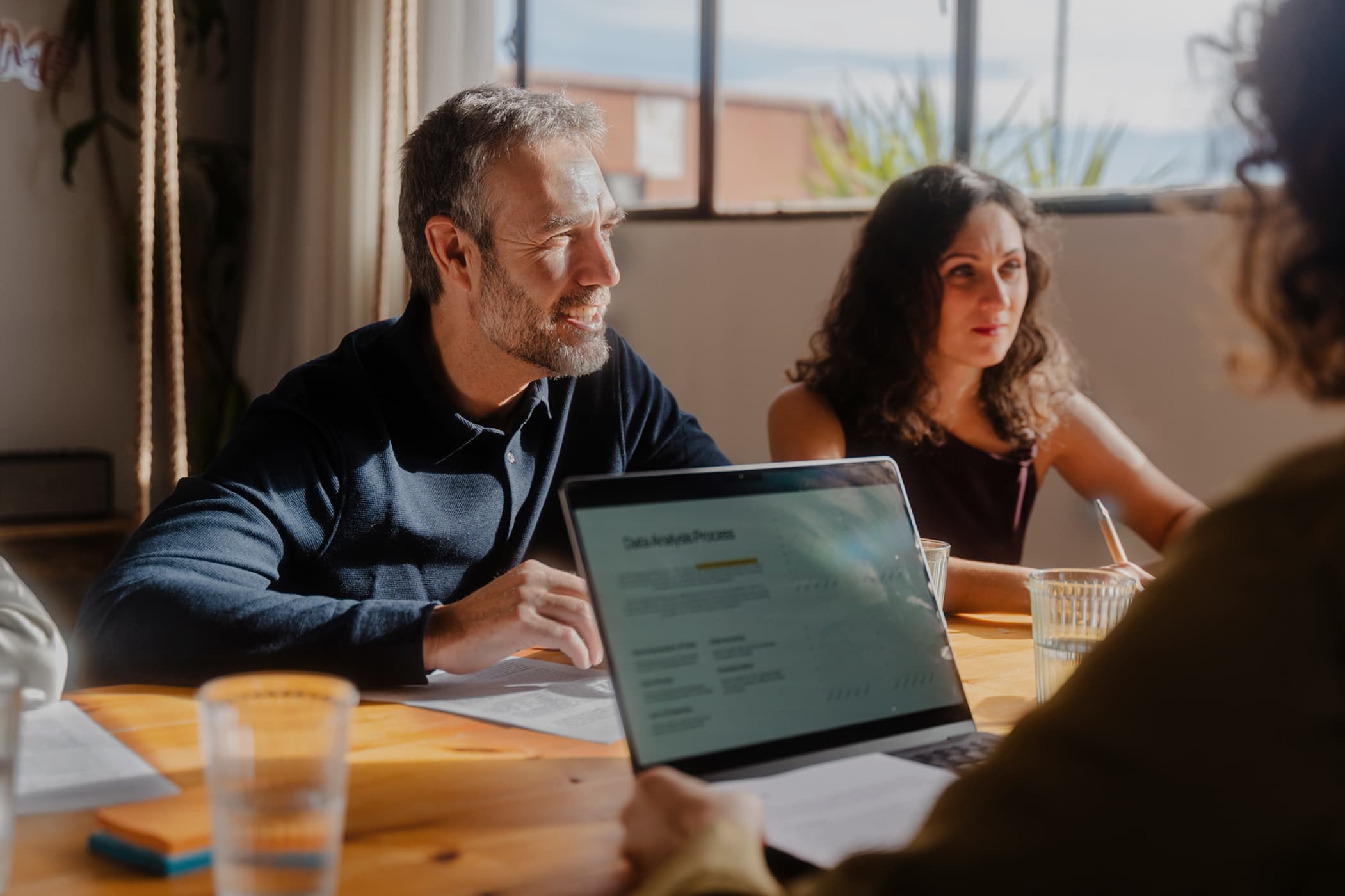 A smiling middle-aged man and a woman listening to a colleague presenting data on a laptop during a meeting.