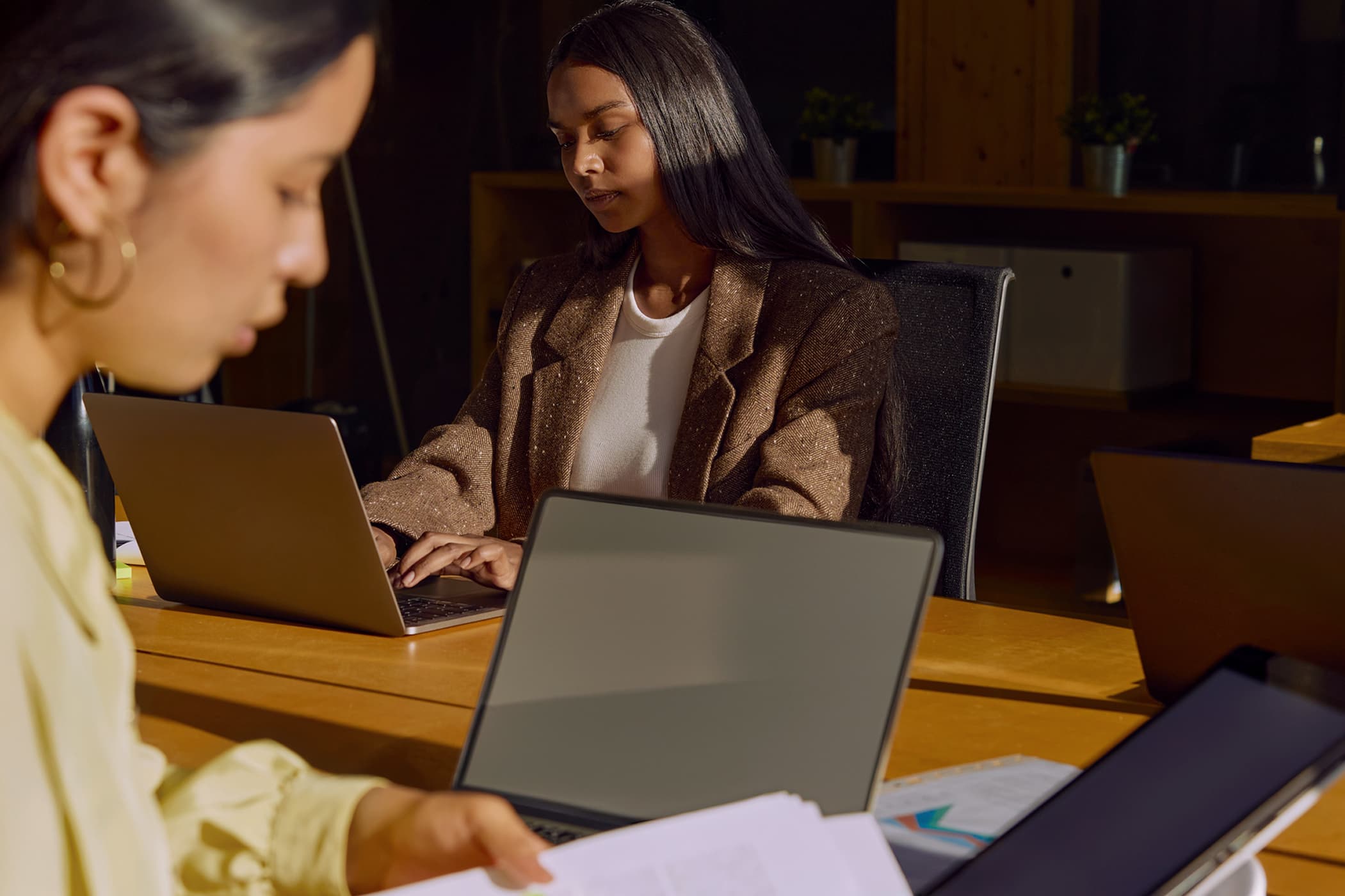 Two women focused on laptops at a wooden desk in a dimly lit office, with one in the foreground reviewing paperwork.