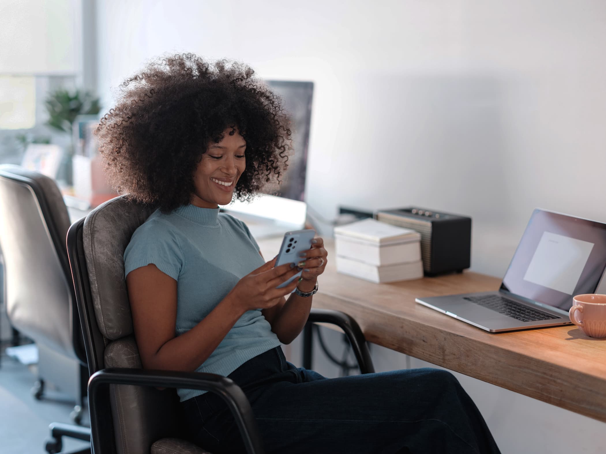 A smiling young woman with curly hair sitting at a wooden desk and looking at her smartphone next to her laptop.