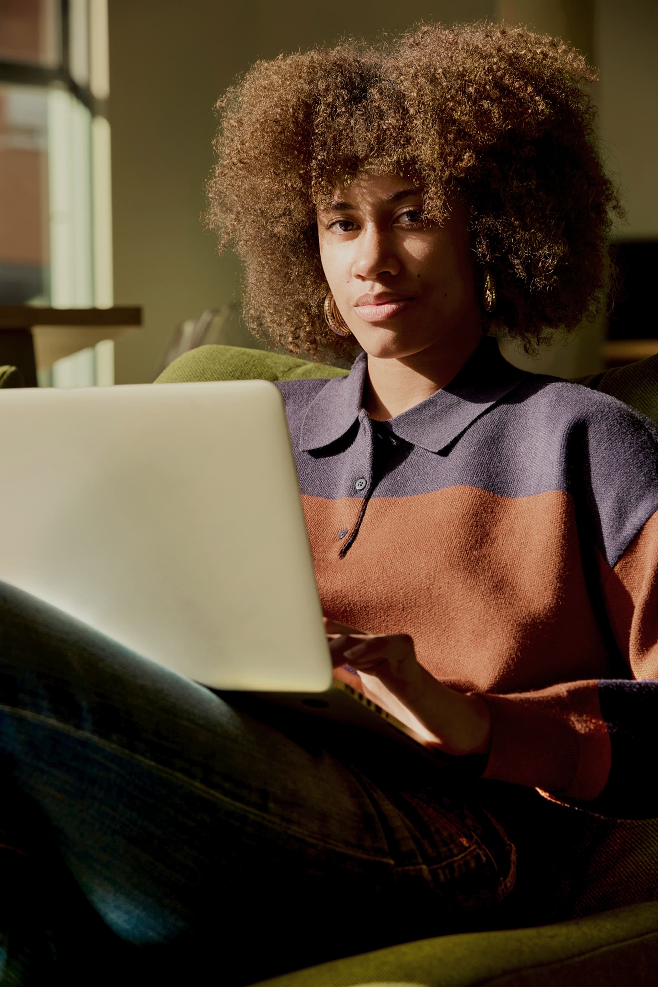 A young woman with afro-textured hair sitting in a chair and typing on a laptop during golden hour.