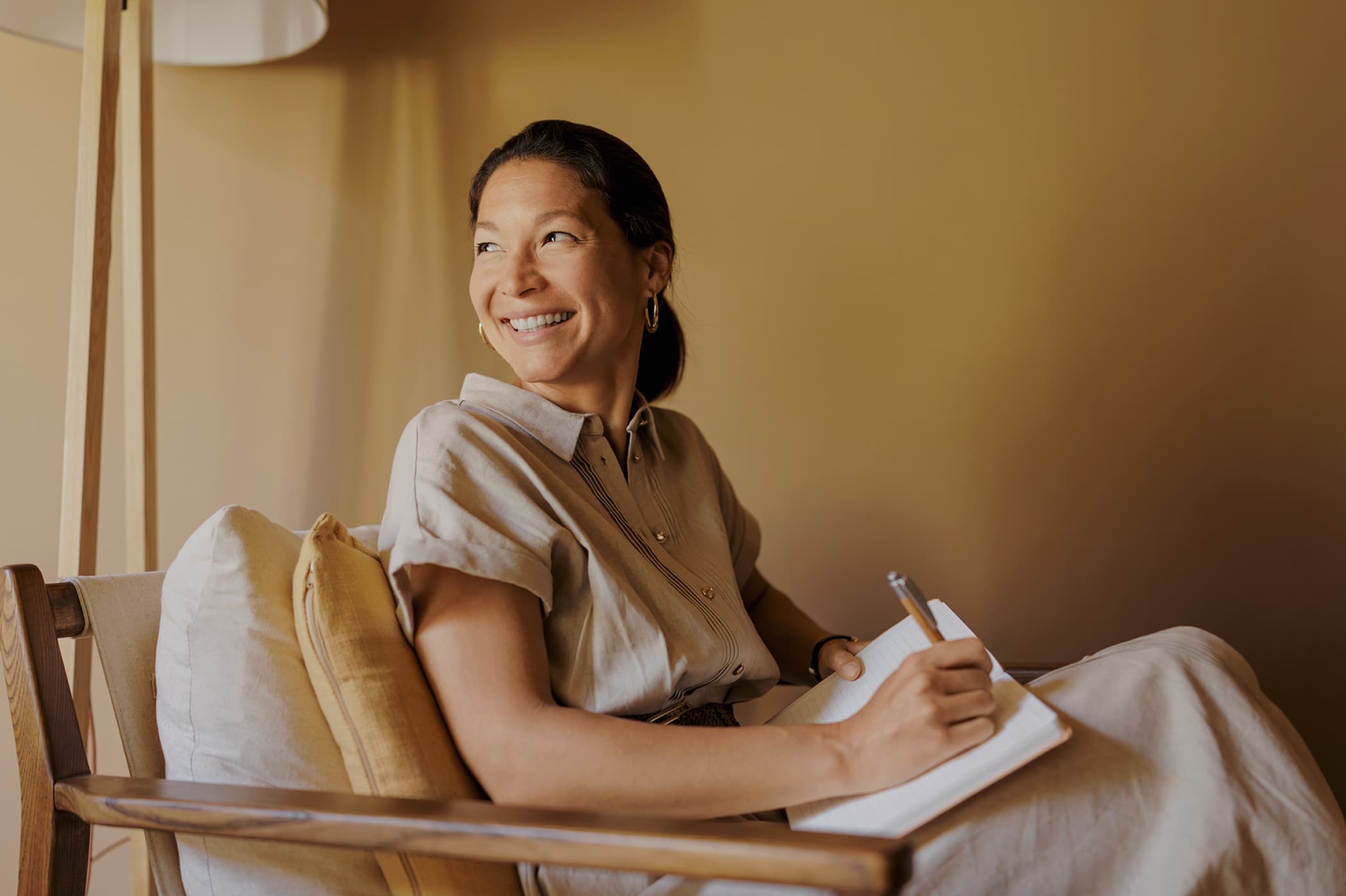 Woman with dark hair sitting in a chair, smiling while writing in a notebook, in a warmly lit room.