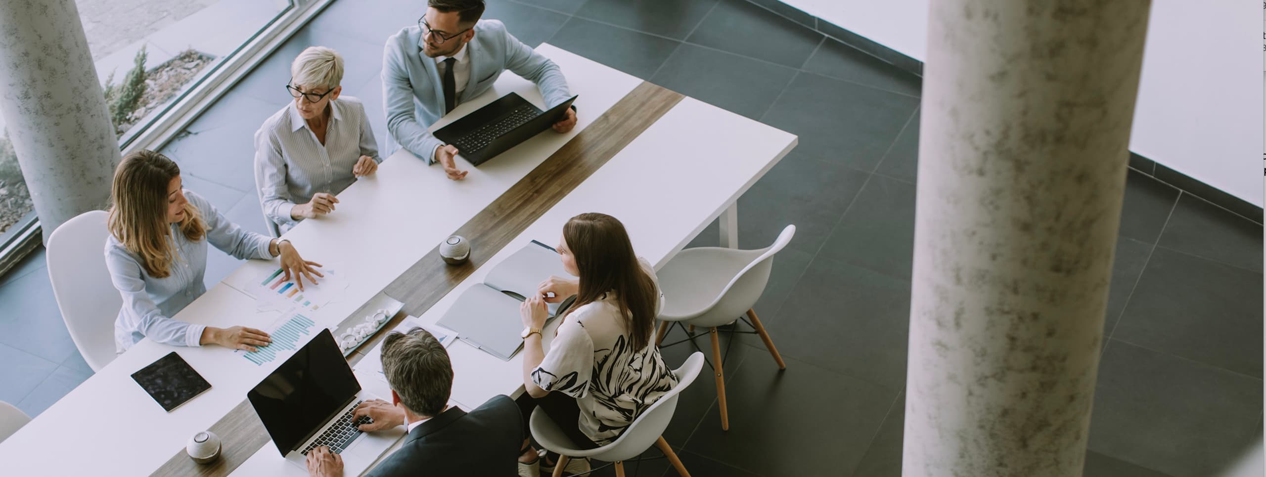 An overhead view of five business colleagues sitting and meeting at a long white conference table in a modern office.