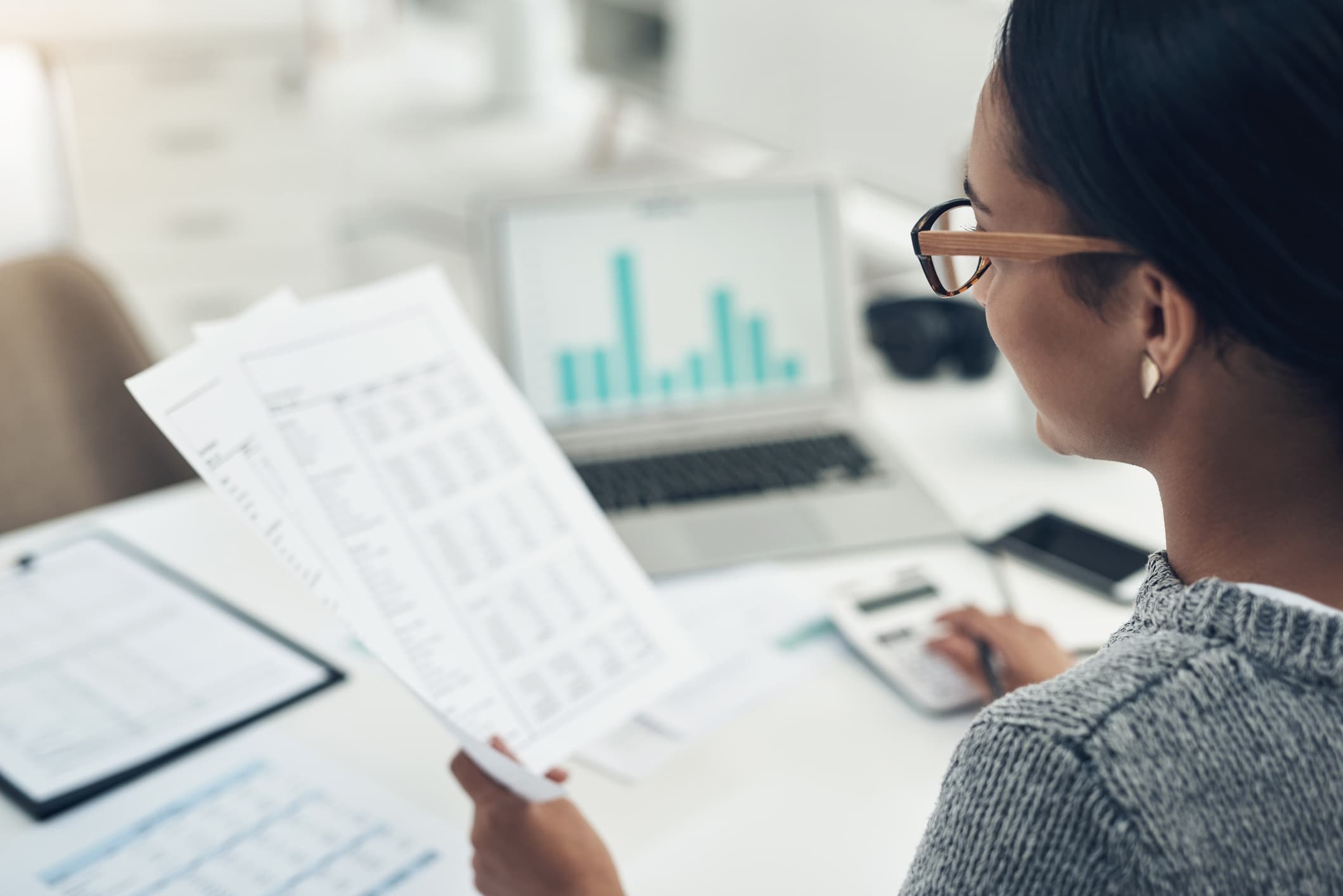 An over-the-shoulder view of a woman with glasses reviewing financial documents at her desk, with a laptop displaying a bar chart in the background.