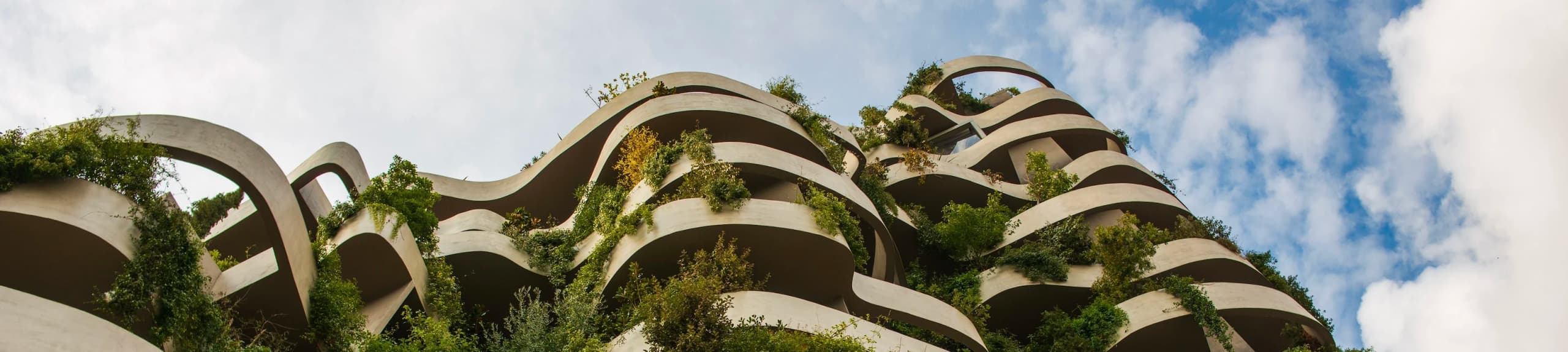 A low-angle view of a modern building with flowing, curved terraces filled with lush green plants against a cloudy sky.