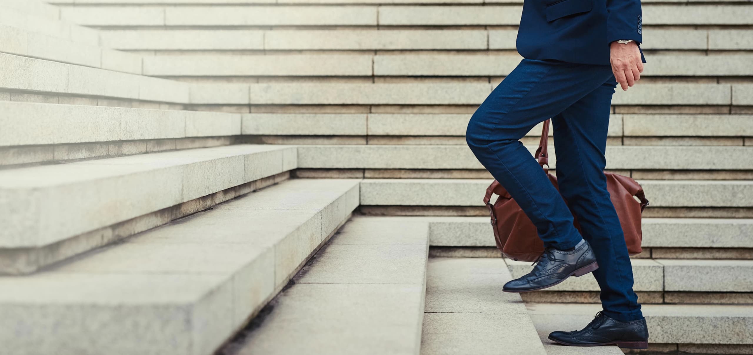A businessman carrying a leather bag, walking up a wide set of outdoor concrete stairs, representing professional growth.