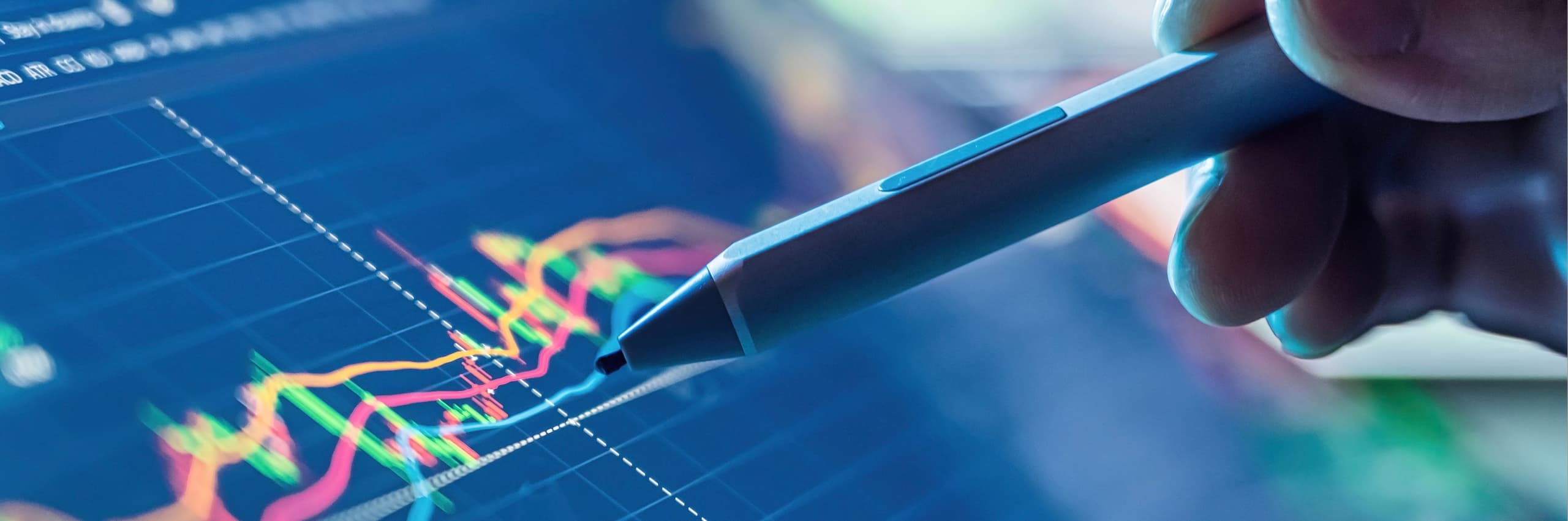 Close-up of a hand pointing a stylus pen at glowing, colorful financial charts and lines of data on a digital tablet.