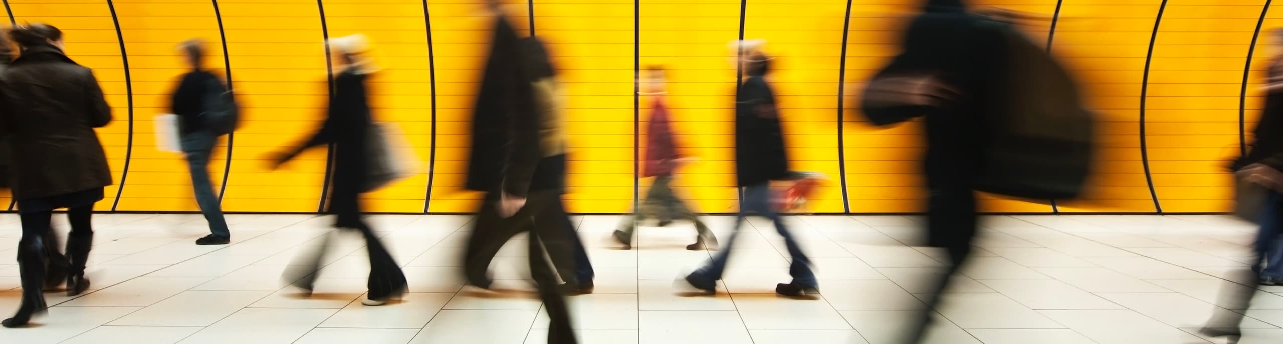 Motion-blurred silhouettes of commuters walking down a bright yellow hallway in a transit station.