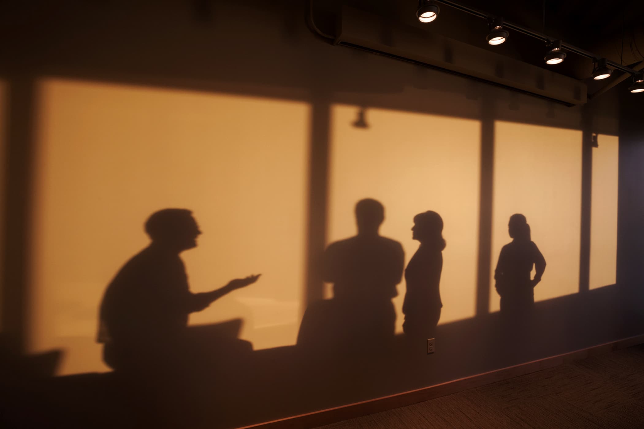 Silhouettes of four people in conversation, cast in shadow against a warmly lit wall in an exhibition space.