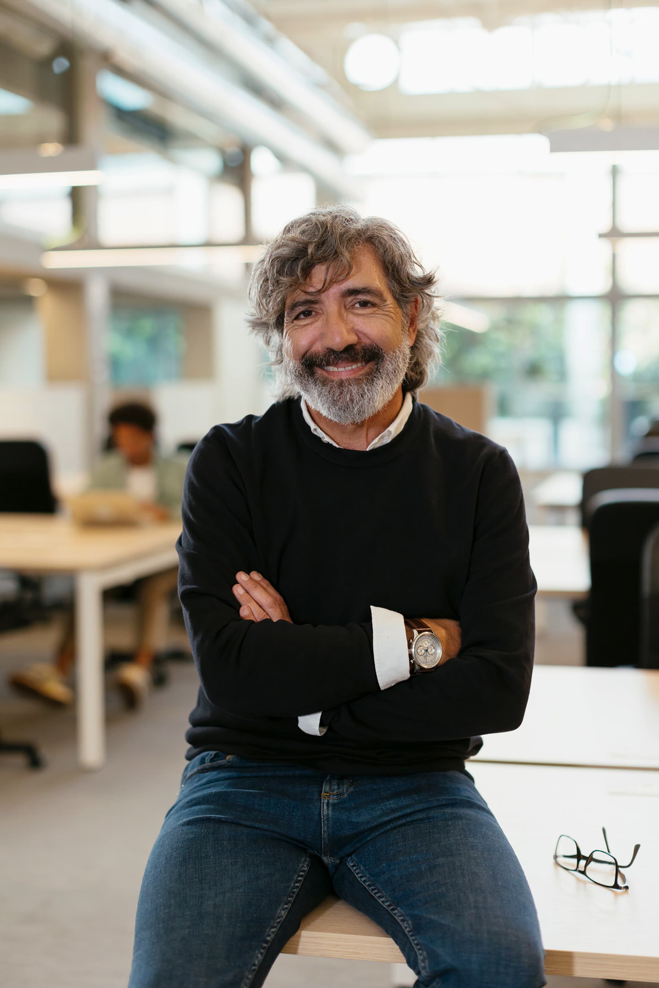 A smiling middle-aged man with wavy grey hair and a beard sitting on a desk with arms crossed in an office.