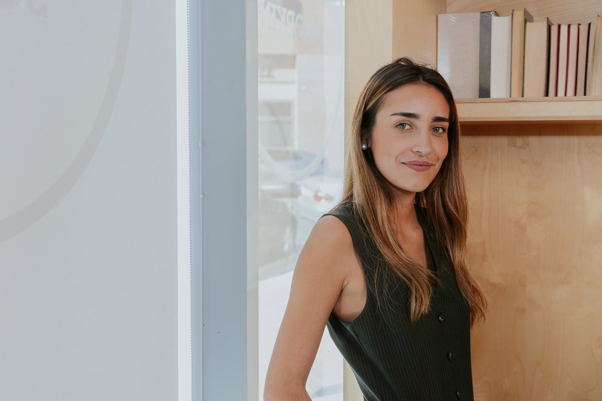 A young woman with long brown hair wearing a dark green sleeveless top, smiling next to a wooden bookshelf.