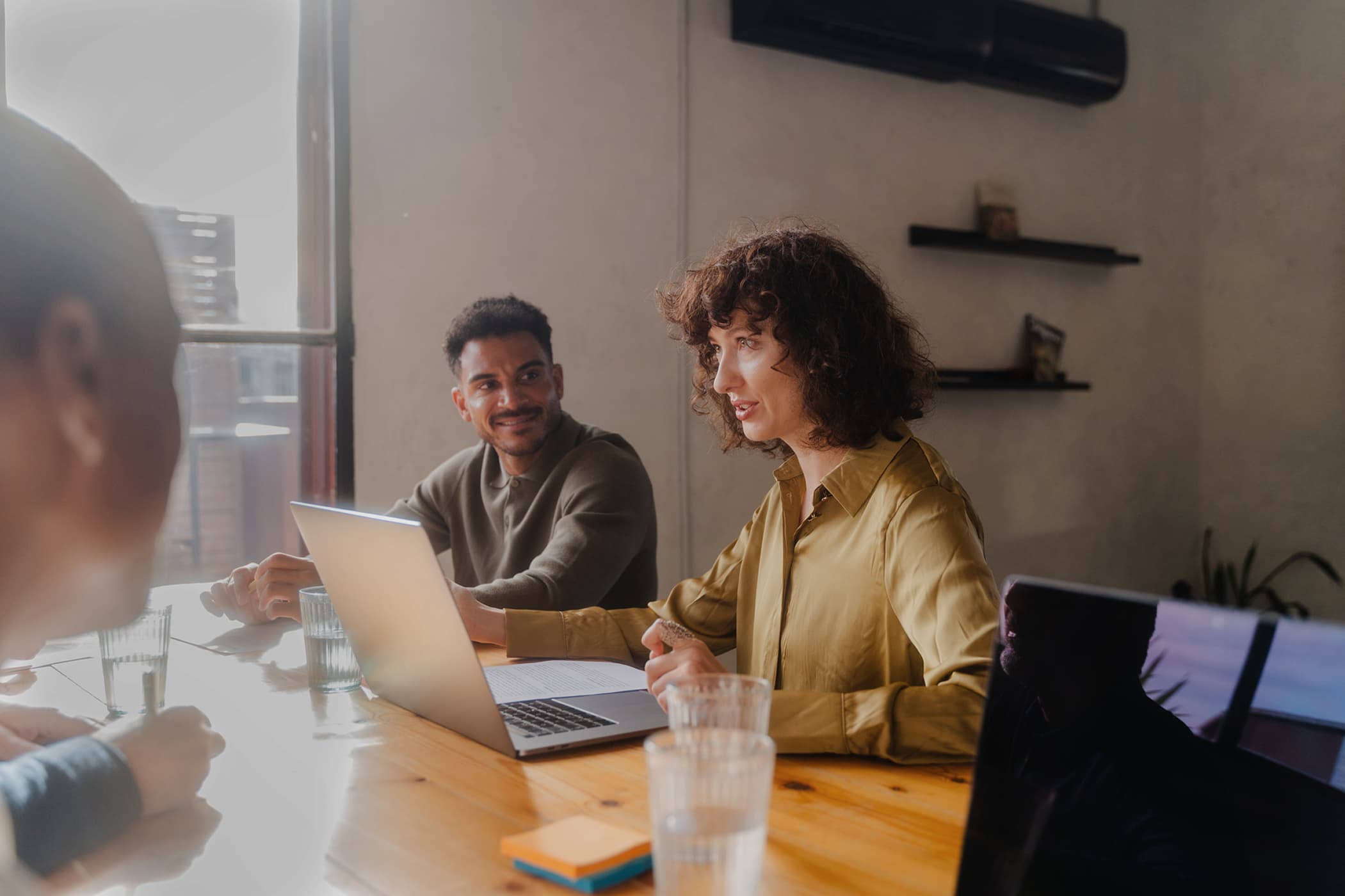 A woman with curly hair speaking during a meeting while her male colleague listens beside a laptop.