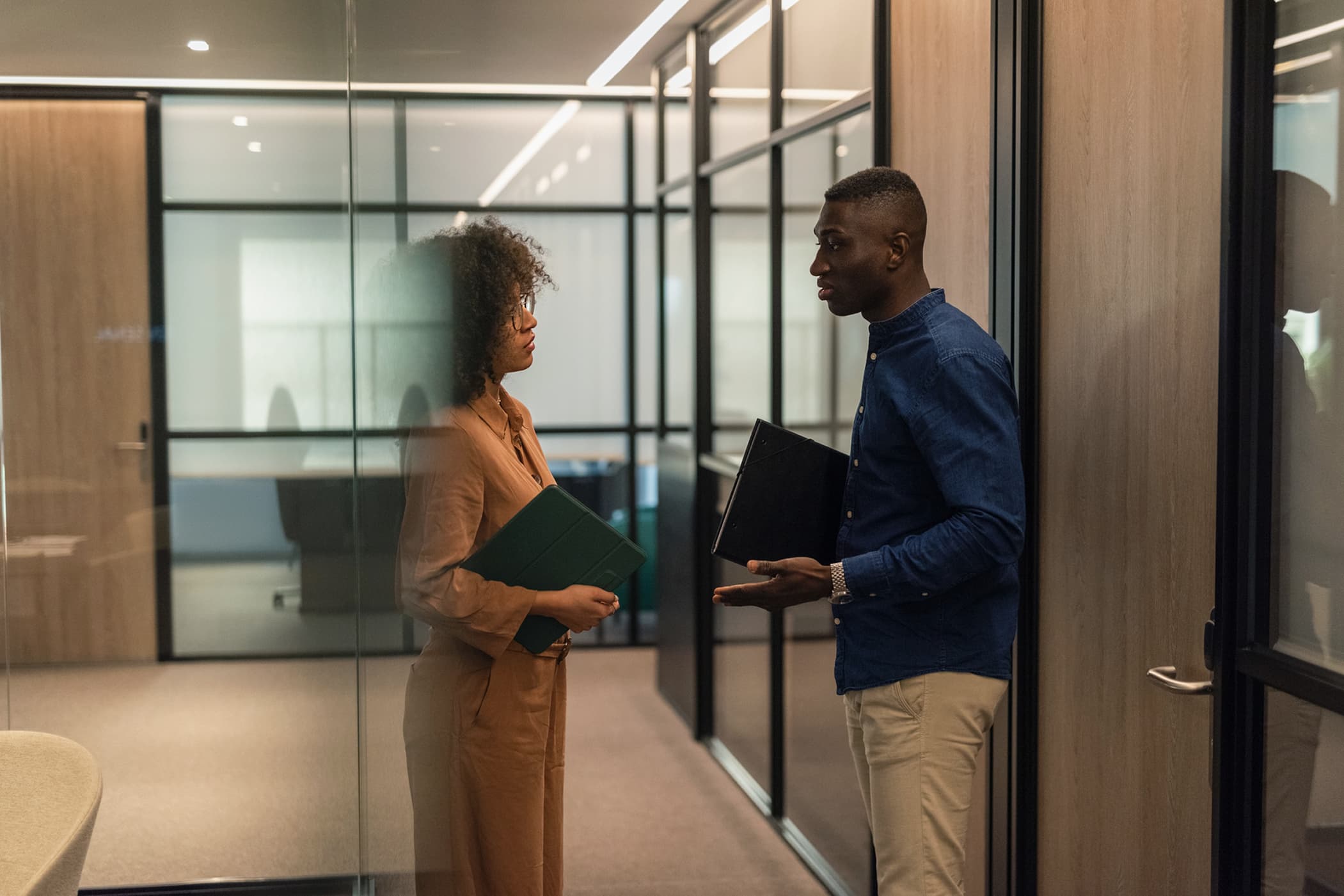 A man and a woman holding folders and talking in a modern office hallway with glass walls.