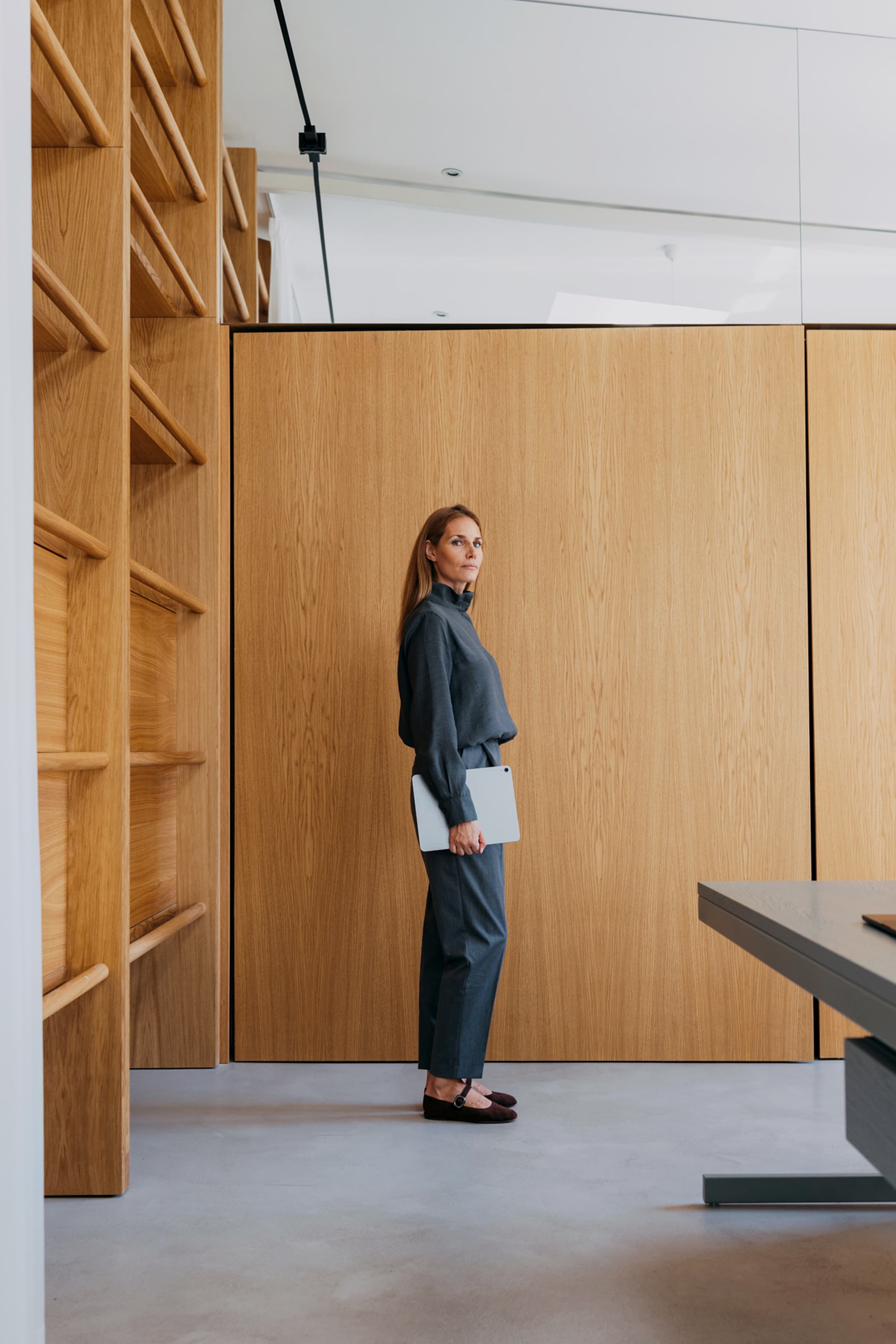 A woman in a dark grey outfit standing in profile, holding a laptop in front of a light wood-paneled wall.