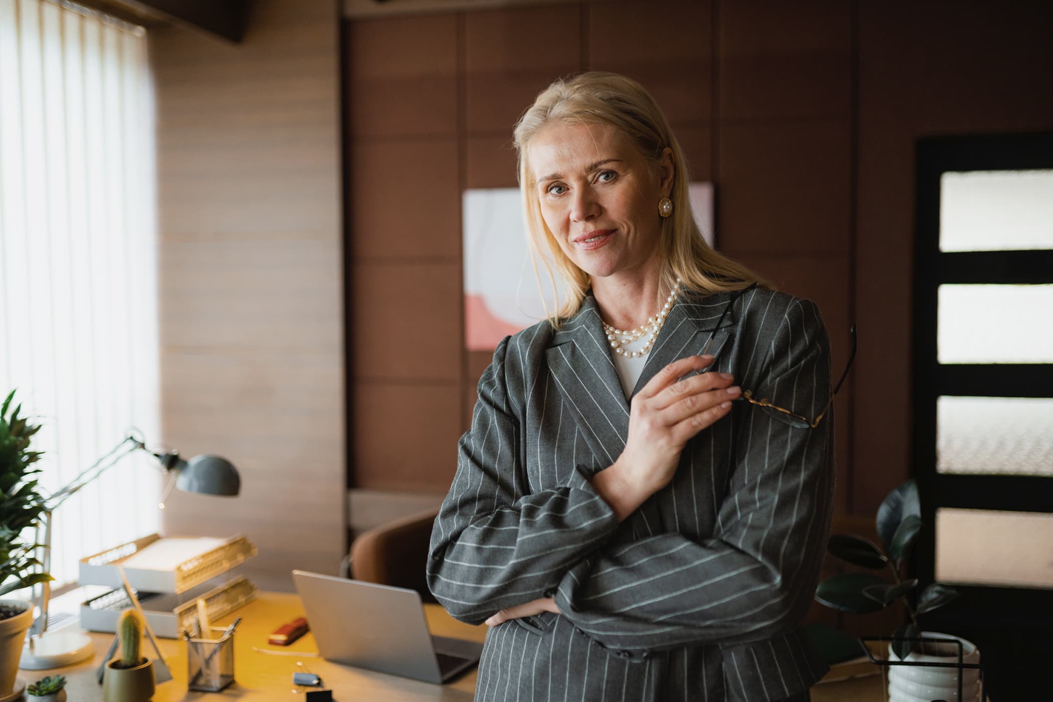 A confident blonde businesswoman in a grey pinstripe suit standing with crossed arms in her office.