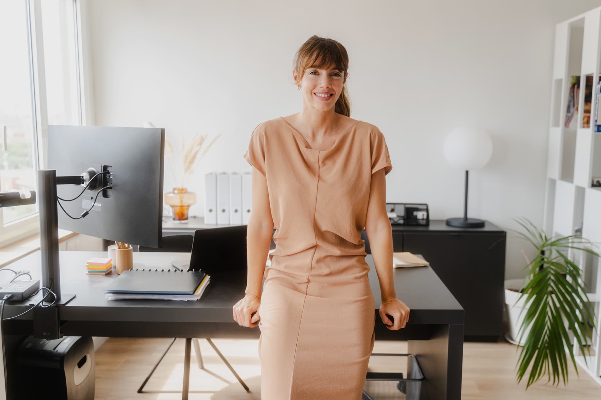 A smiling woman with bangs and a beige dress leaning against a black desk in a bright, modern office.