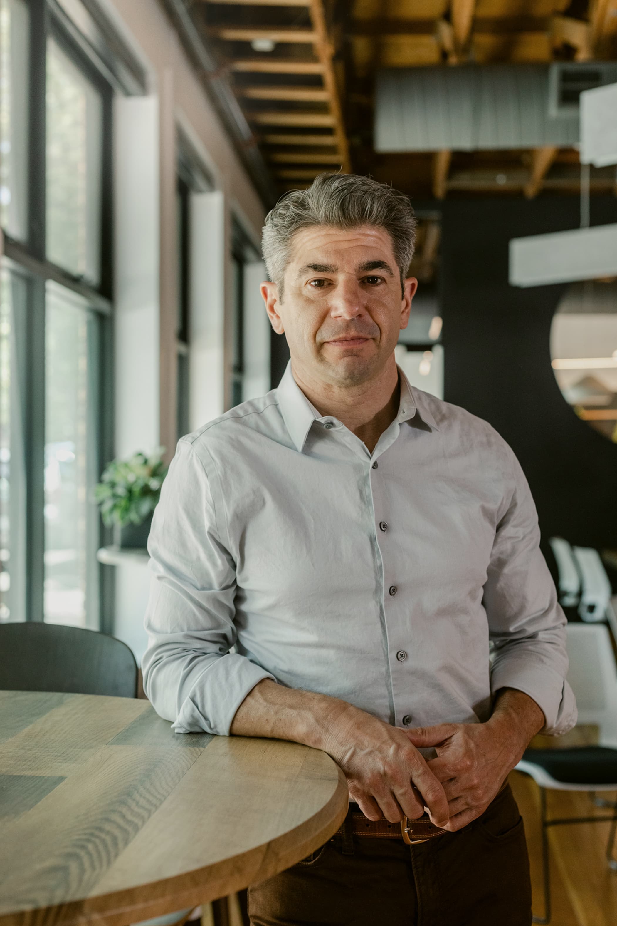 A middle-aged man with grey hair and a light blue button-down shirt leaning against a round wooden table in an office.