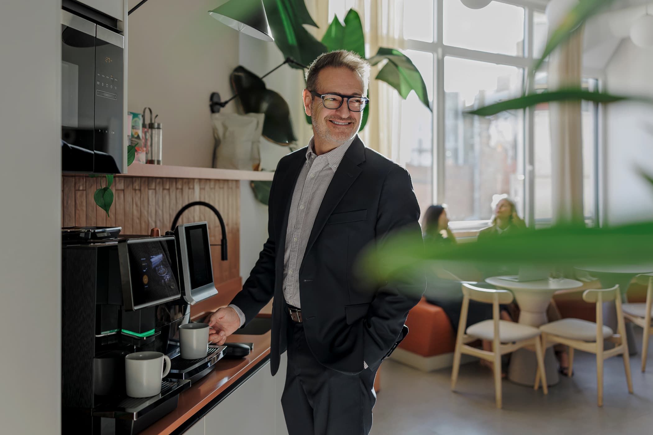 A smiling man in a blazer making coffee at an espresso machine in a modern office kitchen breakroom.